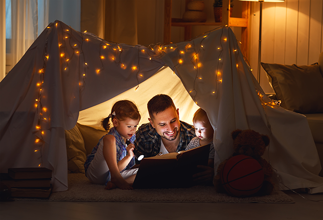 girls and father in a reading tent lit by fairy lights and flashlights.